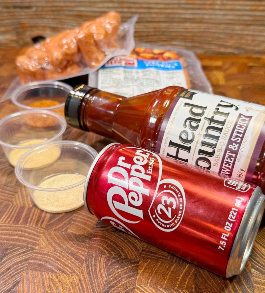 A can of Dr Pepper, a bottle of Head Country Bar-B-Q Sauce, and four small containers of spices are on a wooden surface. In the background, packaged sausages are visible - all ingredients for this lil smokies recipe