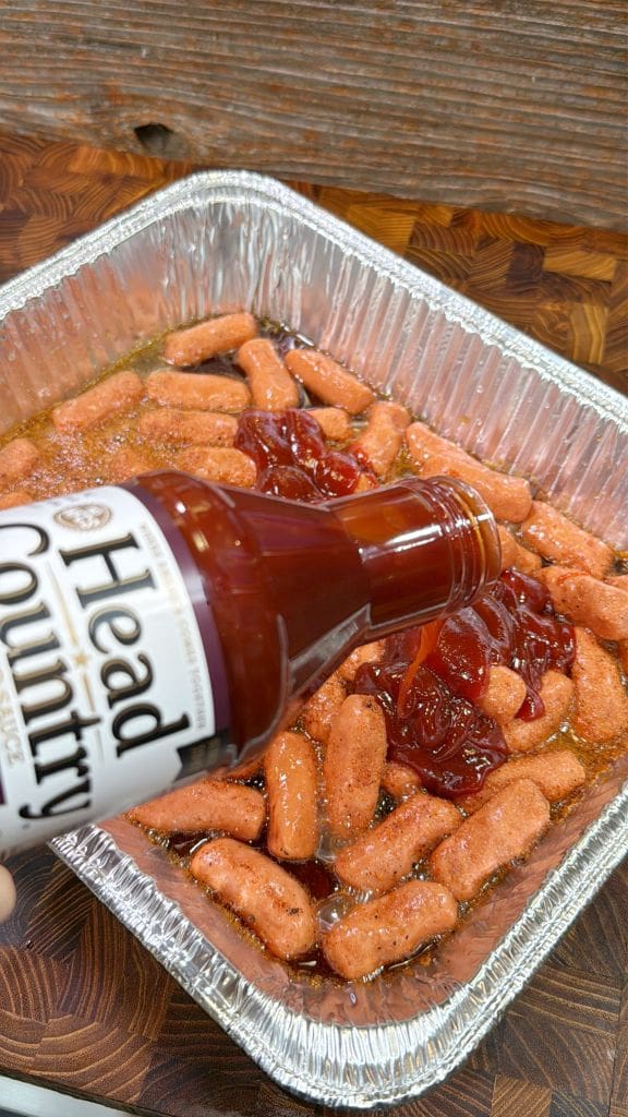 A hand pours barbecue sauce from a Head Country bottle over a foil pan filled with small sausages. The sausages are coated with red sauce and set on a wooden surface.