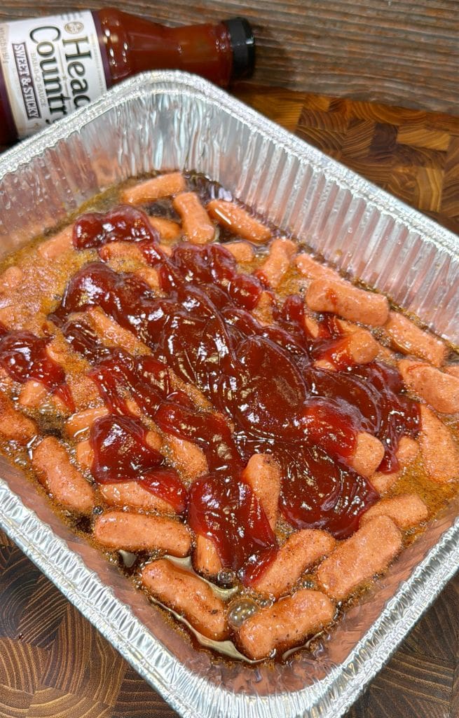 A foil tray filled with mini sausages coated in barbecue sauce, with a bottle of Head Country Bar-B-Q Sauce in the background on a wooden surface.
