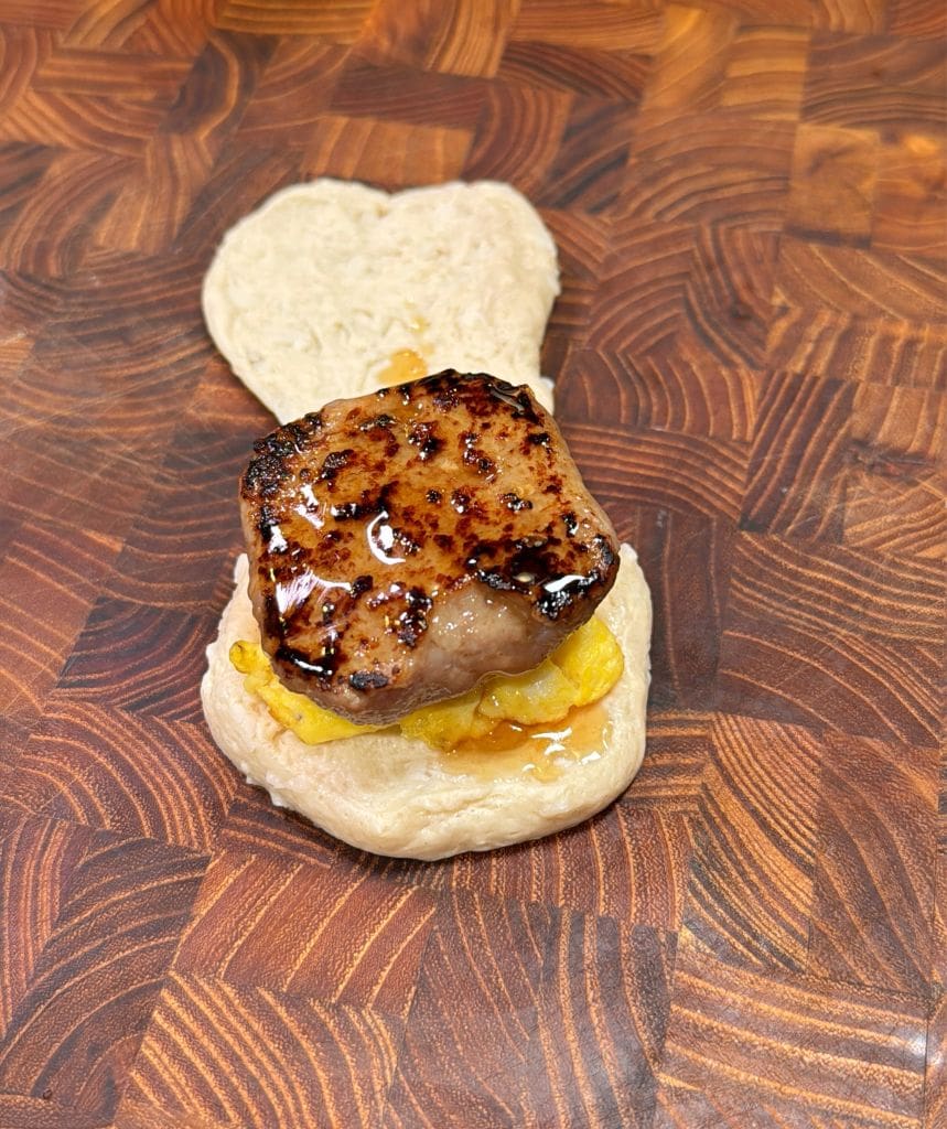 A small sandwich on a wooden surface, featuring a seared piece of meat, possibly lamb or beef, covered with a glossy glaze, and resting on a slice of bread. Theres a yellow condiment or topping underneath the meat.