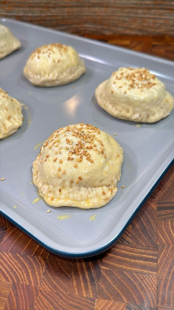 Golden brown, round pastries topped with sesame seeds on a baking tray. The surface is shiny, indicating a glaze. The tray rests on a wooden surface with a textured pattern.