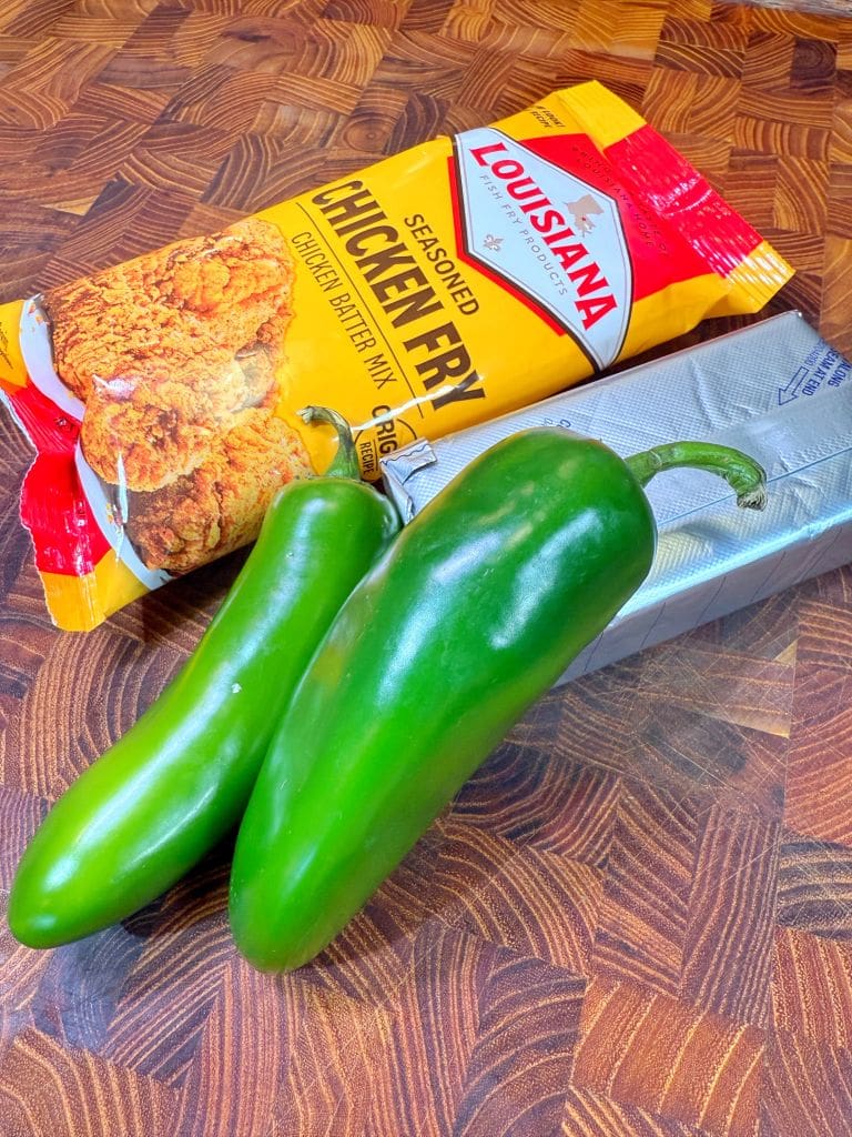 Two green jalapeños sit on a wooden surface alongside a packet of seasoned chicken fry mix and a stick of butter. The packet has Louisiana branding visible.
