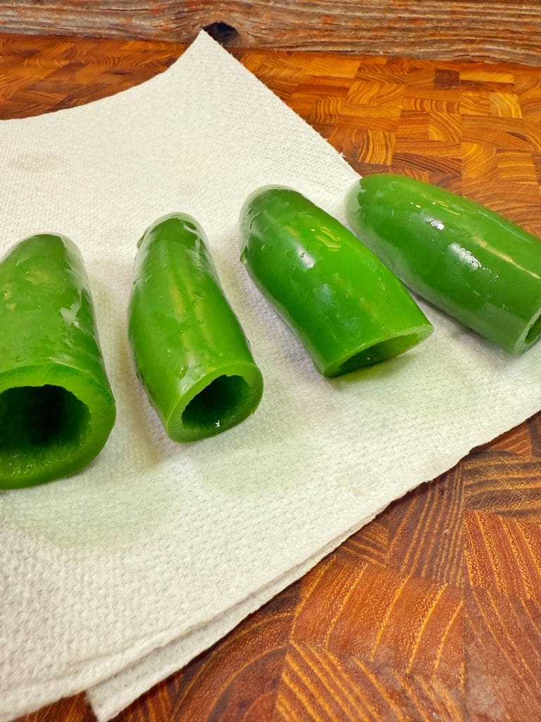 Four hollowed-out green jalapeño peppers are placed in a row on a white paper towel. The paper towel rests on a wooden surface with a textured pattern. The peppers are shiny and fresh.