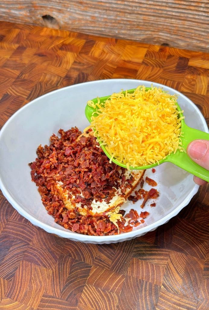 A bowl on a wooden surface contains chopped bacon and cream cheese. A hand is adding shredded cheddar cheese from a green measuring cup into the bowl.