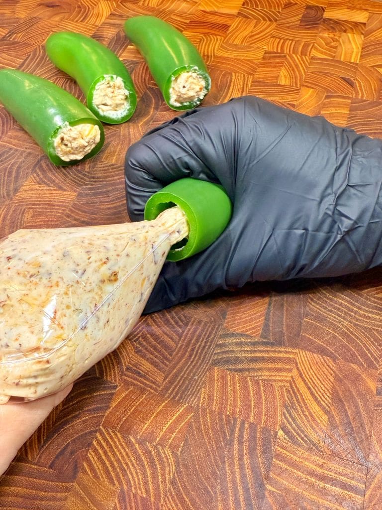 A hand wearing a black glove stuffs a green jalapeño pepper with a mixture using a piping bag. In this chicken fried recipe three stuffed jalapeños are visible in the background, placed on a wooden surface.