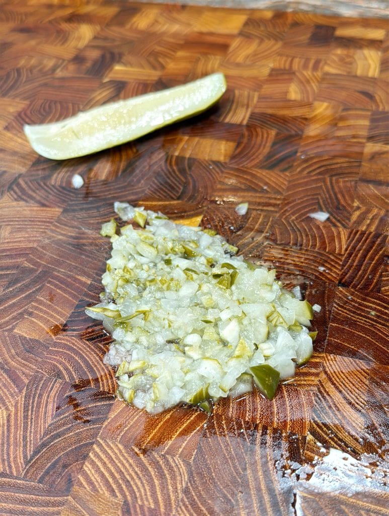 Chopped pickles on a wooden cutting board with a pickle wedge in the background. The board has a detailed, patterned surface.