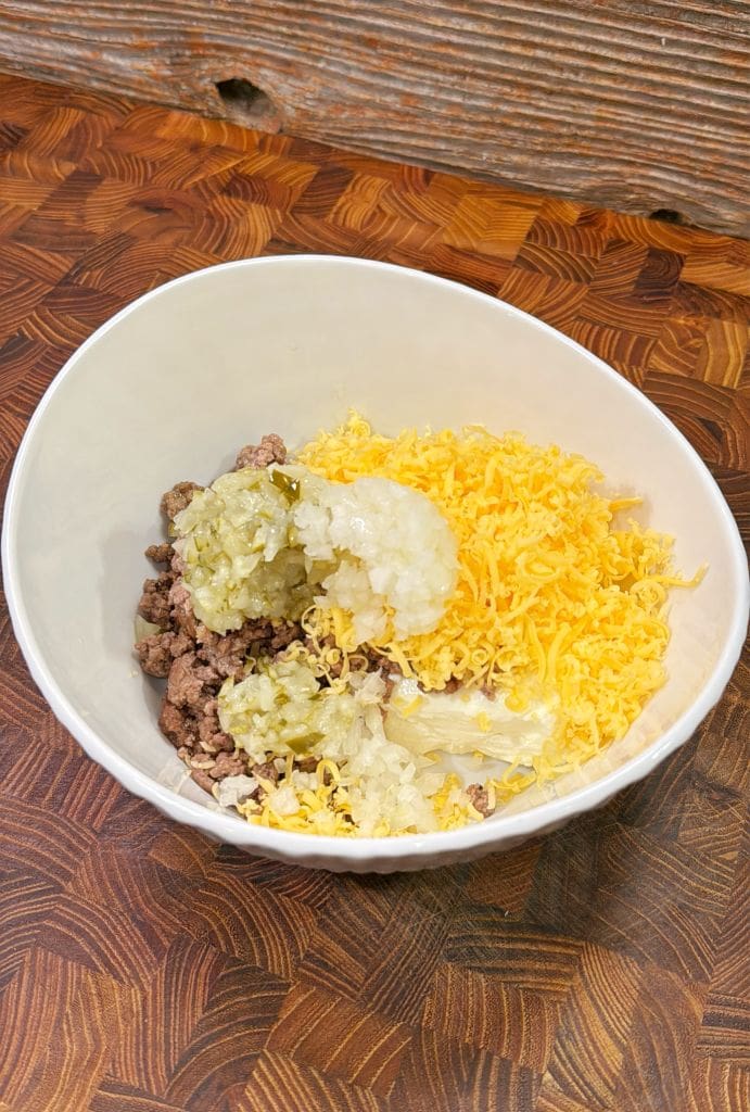 A bowl filled with cooked ground meat, shredded cheddar cheese, and a topping of pickles with mayonnaise or a similar dressing, all placed on a wooden countertop.