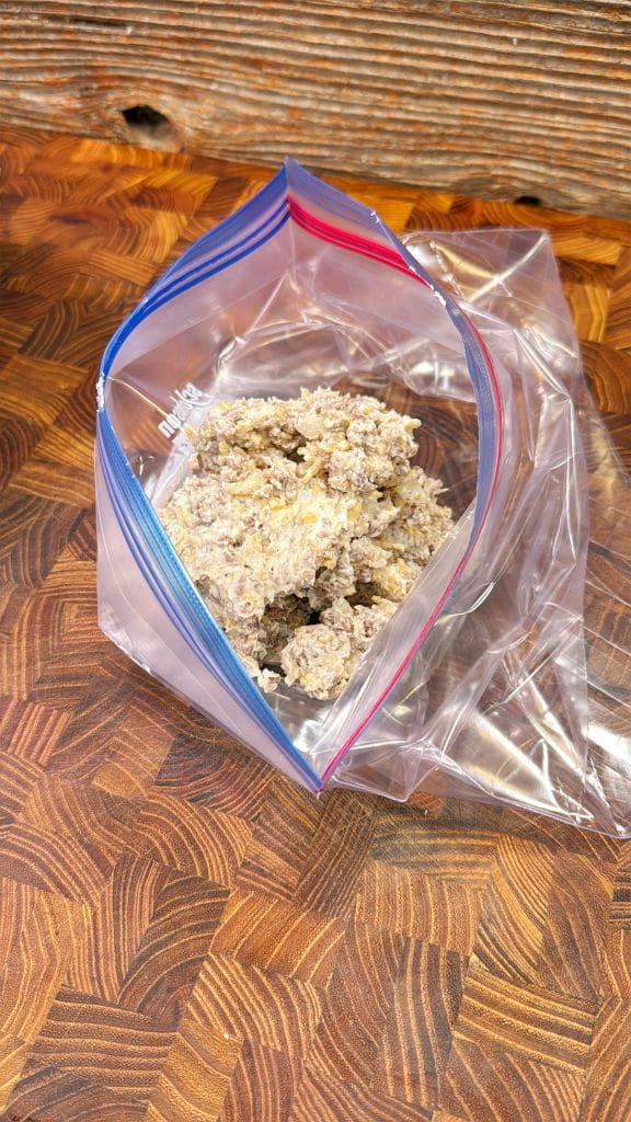 A plastic zipper bag partially filled with a crumbly, beige mixture, possibly food, resting on a wooden surface with a textured, geometric pattern. The bag has a blue and red seal.