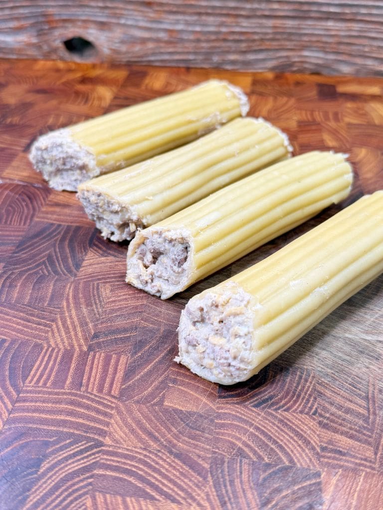 Four uncooked cannelloni shells filled with a white meat mixture are arranged on a wooden cutting board with a geometric pattern. The background features a textured wooden surface.