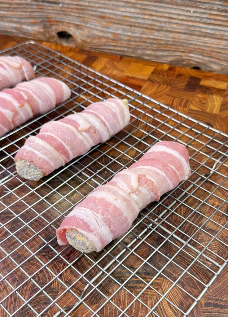 Uncooked sausage rolls wrapped in bacon are placed on a wire rack over a wooden surface, ready for cooking.
