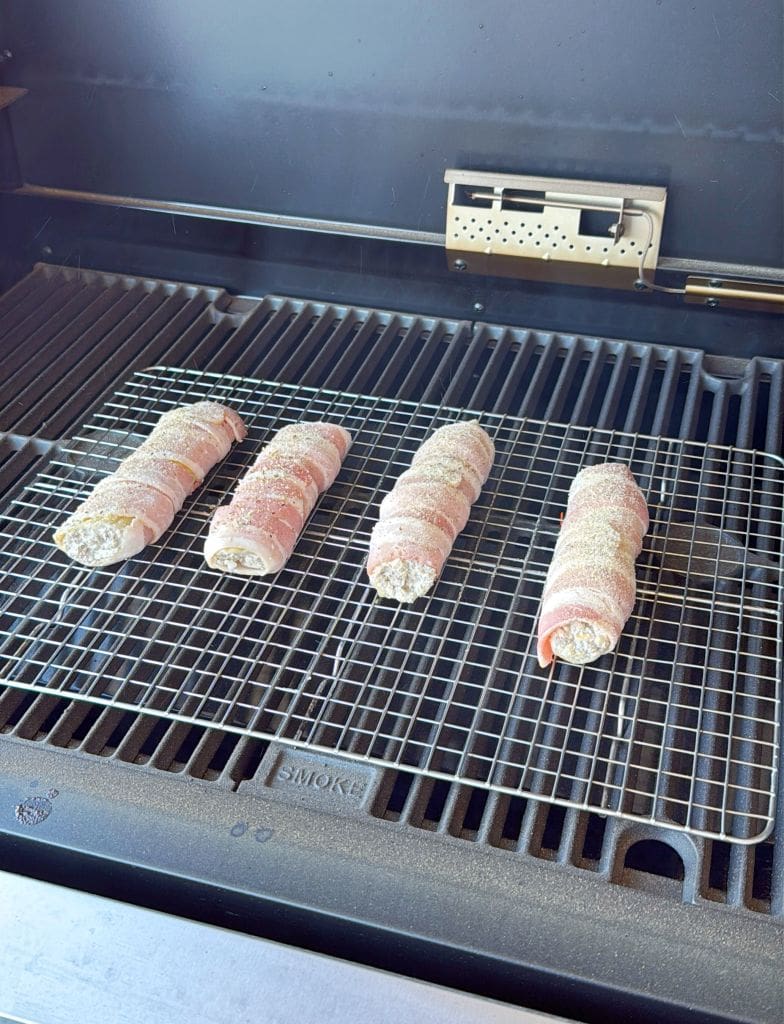 Four bacon-wrapped items on a wire rack inside a grill. The grill grate is visible underneath, and the items appear evenly spaced. The background shows part of the grill interior.