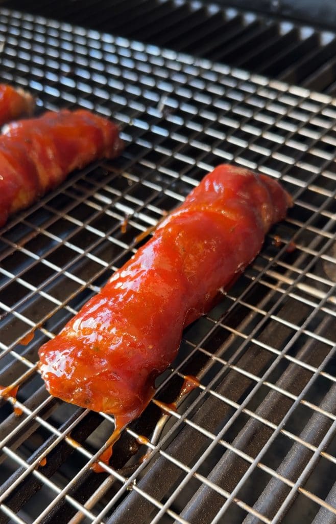 Shotgun Shells recipe: bacon wrapped manicotti wrapped in  bacon, is coated in a glossy reddish-brown sauce as it rests on a metal grill. The sauce glistens under the light, showcasing the grills grid pattern beneath this tempting culinary piece.