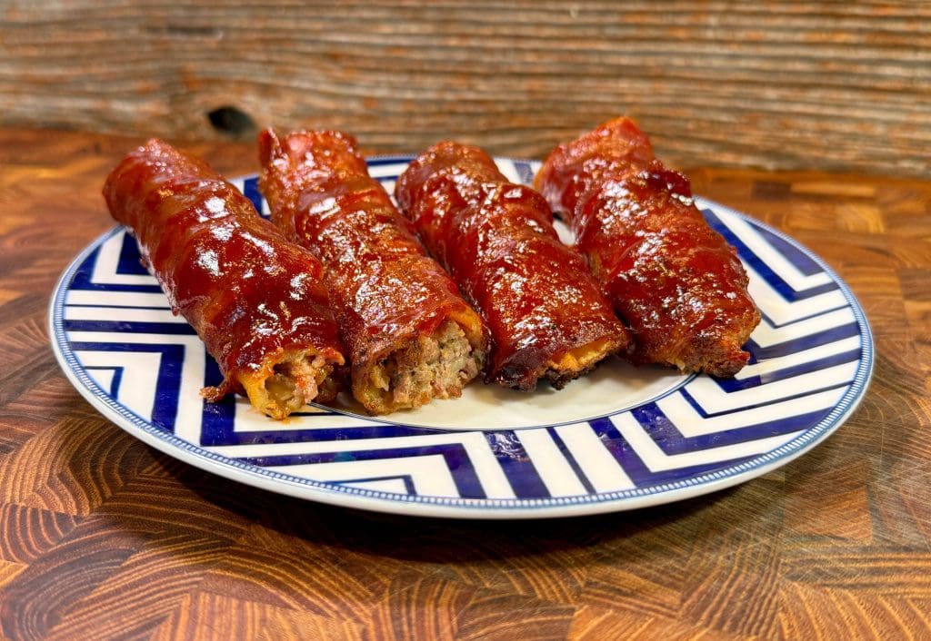 Four bacon-wrapped meatloafs, reminiscent of decadent cheeseburgers, are glazed with a shiny sauce and arranged on a blue and white patterned plate. The background features a wooden table surface and a textured wooden wall.