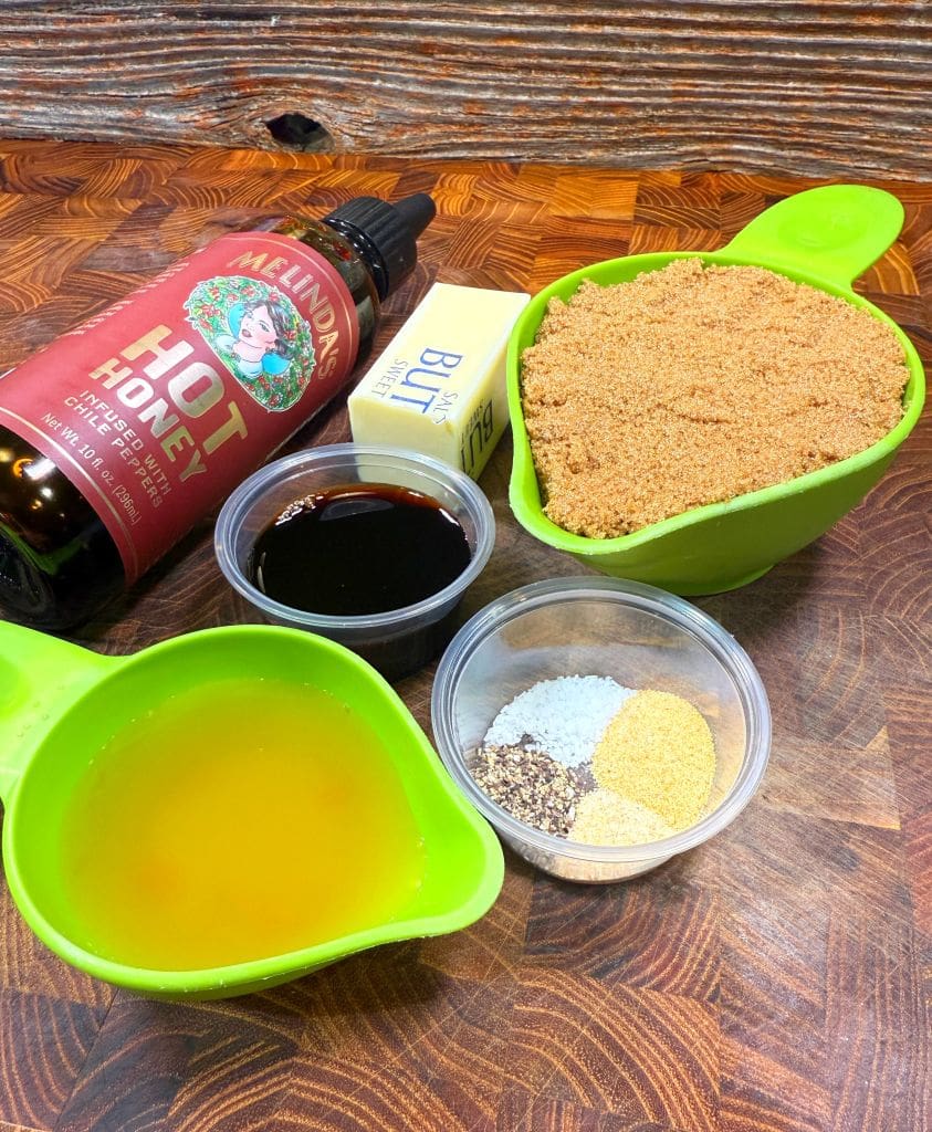 Ingredients for a recipe on a wooden surface: a bottle of hot honey, a stick of butter, a cup of brown sugar, a measuring cup of yellow liquid, a small container of dark liquid, and another with various spices.