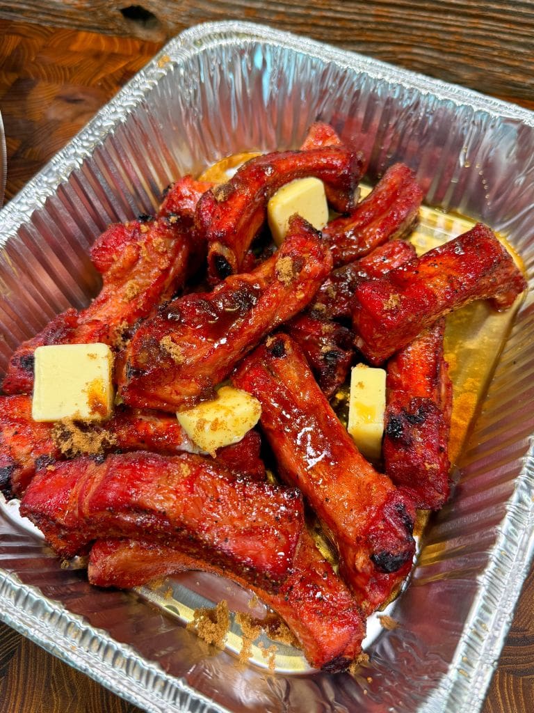 A foil tray filled with glazed, grilled pork ribs topped with pats of butter and sprinkled with brown sugar on a wooden table. The ribs are richly colored and glistening, suggesting a savory and sweet preparation.