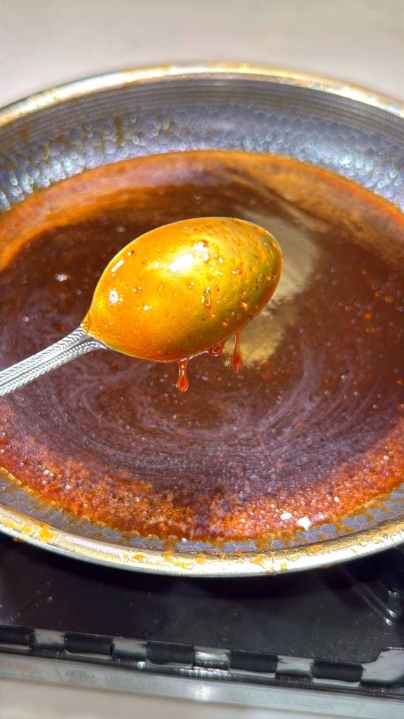 A spoon lifts thick, brown caramel sauce from a metal pan. The sauce glistens and drips slightly from the spoon, with a golden sheen on its surface. The pan beneath is coated with a thin layer of the same caramel.