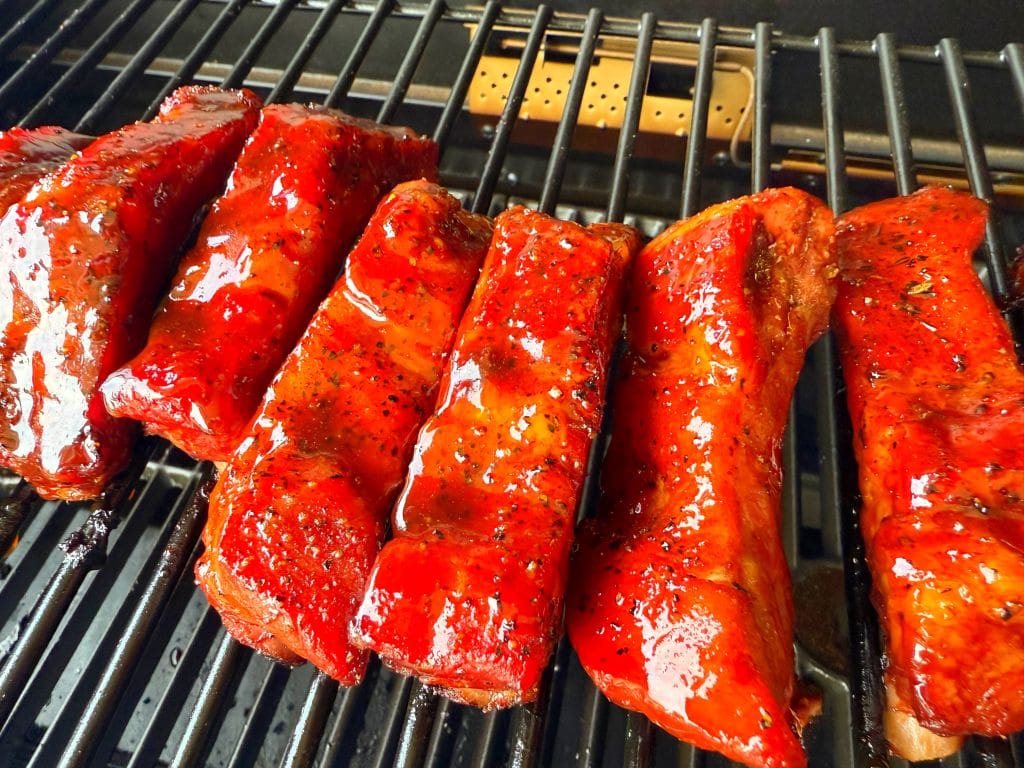 Close-up of glazed barbecue ribs on a grill. The ribs are coated in a shiny, rich red sauce, and are placed in a row on the metal grill grate, suggesting theyre being cooked or smoked, with a few grill marks visible.