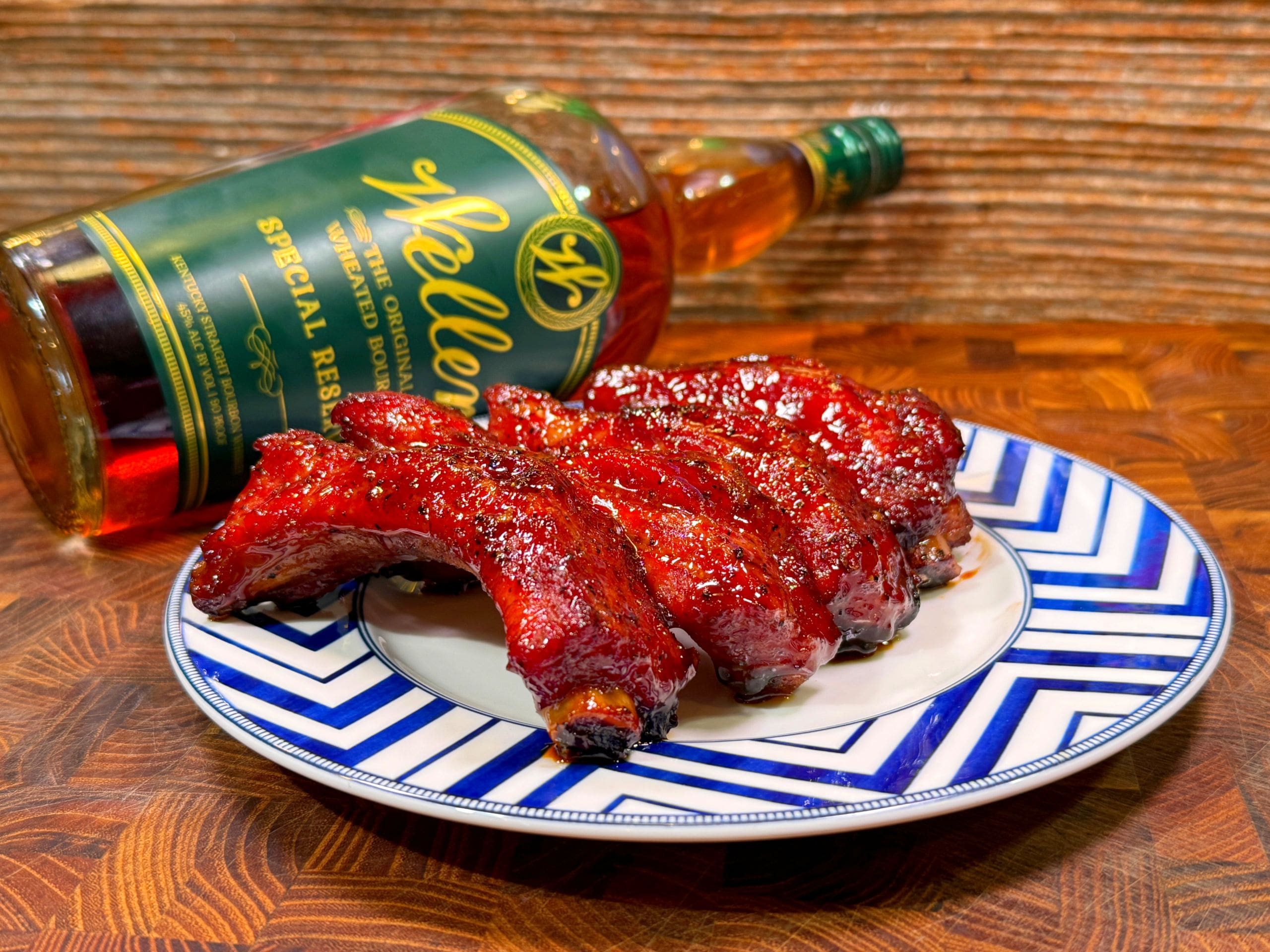 A plate of glazed ribs on a blue and white patterned plate with a bottle of whiskey in the background, set on a wooden table.