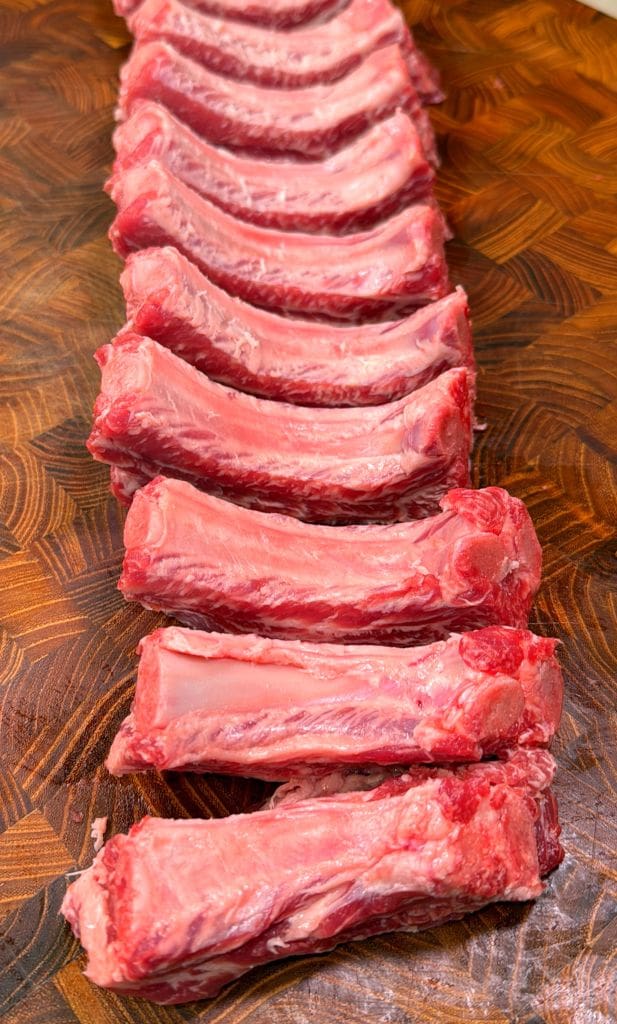 Raw pork ribs for a party ribs recipe neatly arranged in a row on a wooden cutting board. The ribs are pink and marbled with fat, showcasing fresh and uncooked meat with visible bones. The wooden surface has a patterned grain.