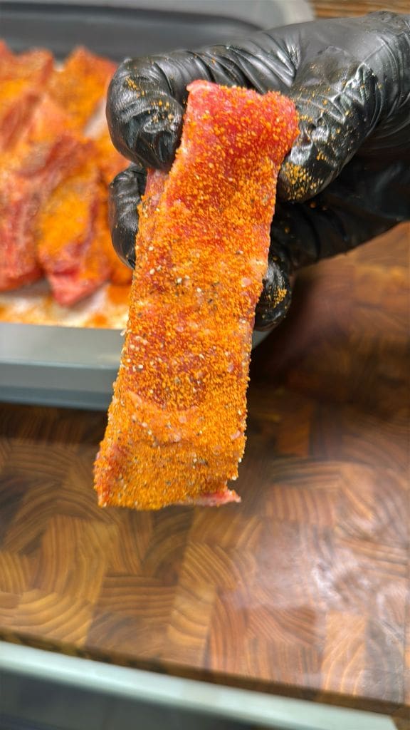 A gloved hand holding a strip of raw meat seasoned with a reddish spice blend, with more seasoned meat visible in a tray in the background.