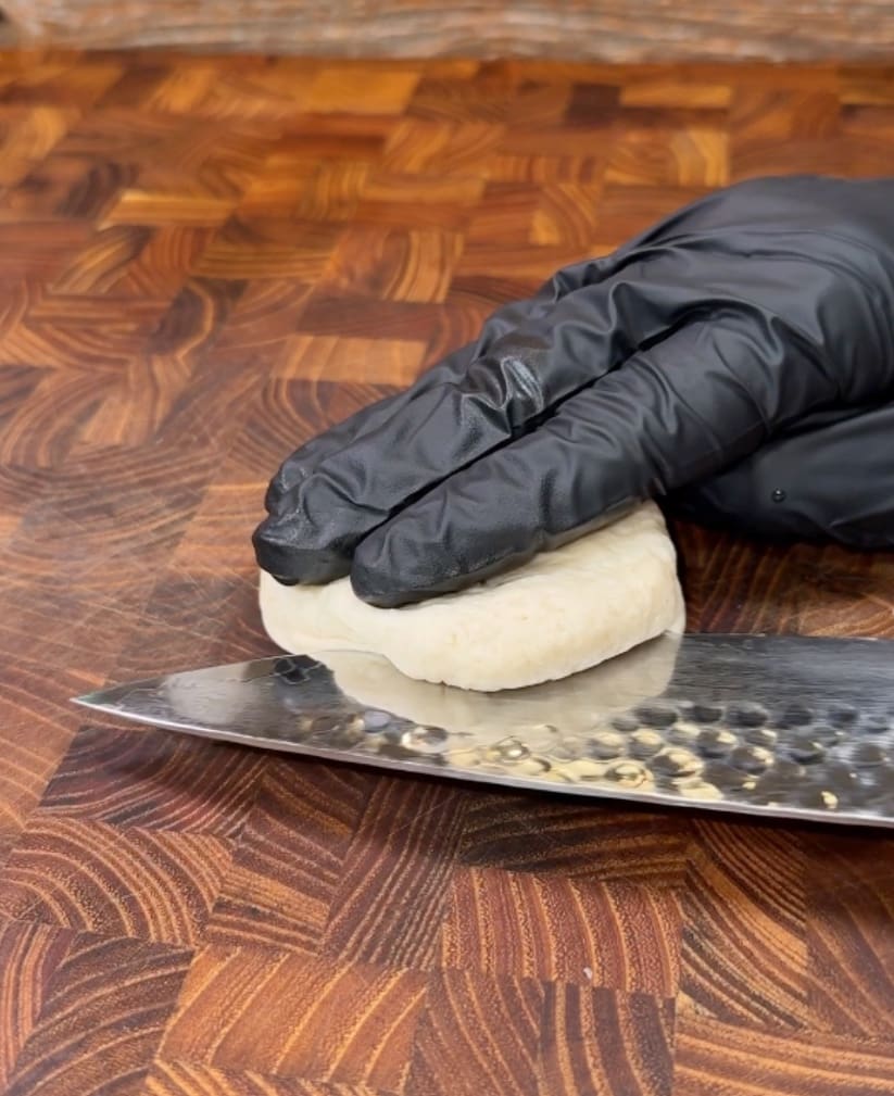 A person wearing a black glove slices a piece of dough with a serrated knife on a wooden cutting board.