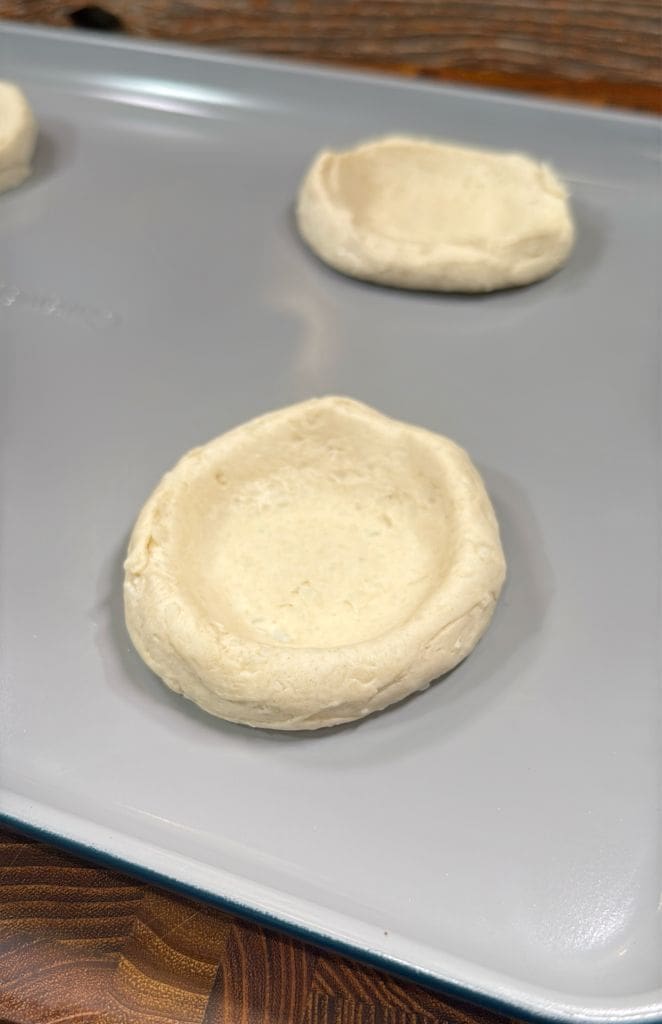 Close-up of unbaked dough rounds with indented centers on a baking sheet, ready for filling.