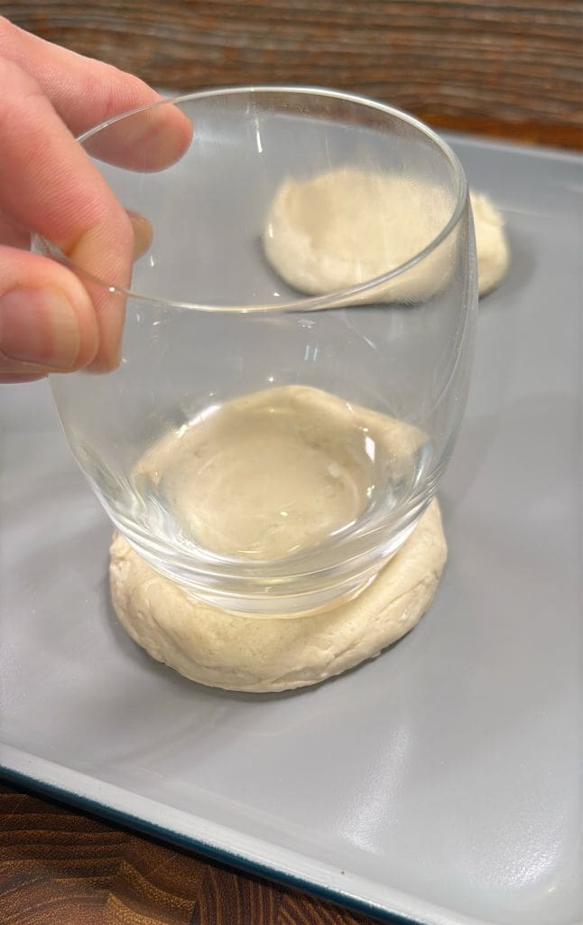 A hand pressing a glass onto a ball of dough on a gray baking sheet, flattening it into a disc. Another dough ball is visible next to it. The surface beneath the baking sheet is wooden.