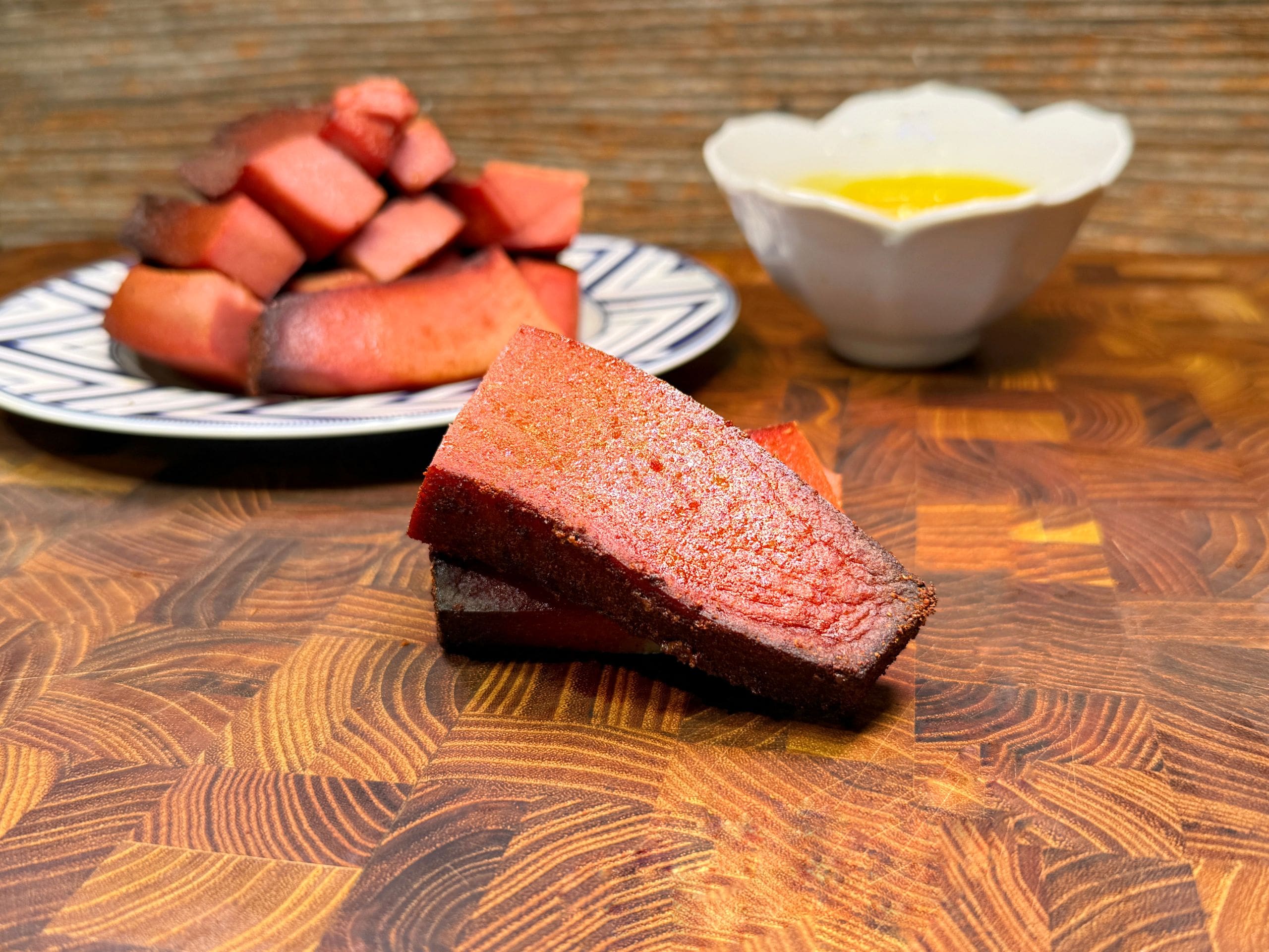 Two slices of smoked watermelon are placed on a wooden board. In the background, a plate with more slices and a bowl of yellow sauce are visible. The setup is on a textured wooden surface.