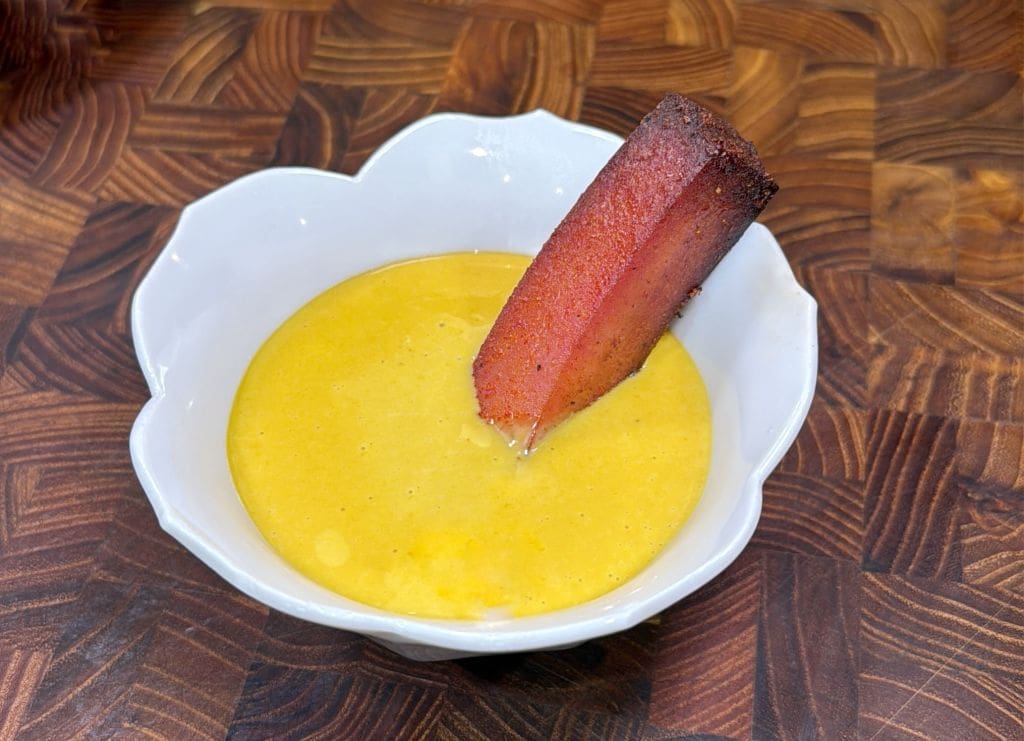 A white bowl filled with creamy yellow honey mustard is placed on a wooden surface. A piece of bologna from the smoked bologna recipe is standing upright.