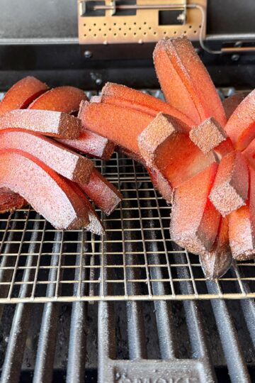 Two reddish-brown slices of Bologna scored and fanned out like blooming onions rest on a grill rack inside a smoker grill. The edges are crisp, revealing a textured surface, with smoke marks visible underneath.