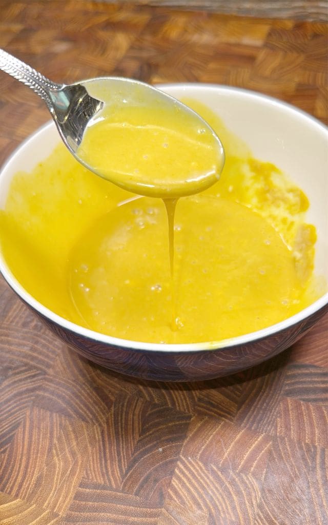 A spoon drizzling a thick, yellow liquid back into a white bowl on a wooden surface, creating a smooth texture. The liquid appears to be a sauce or dressing.