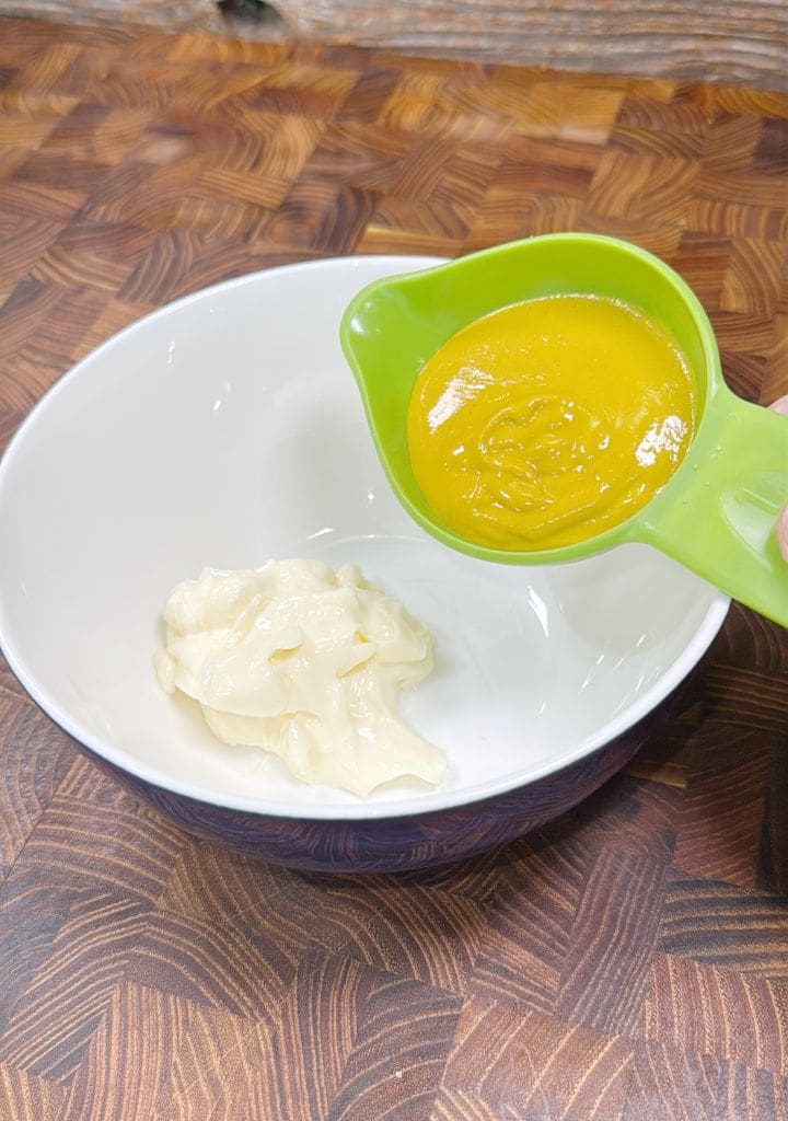 A green measuring cup is pouring yellow mustard into a white mixing bowl containing mayonnaise. The bowl is placed on a wooden countertop with a geometric patterned surface.
