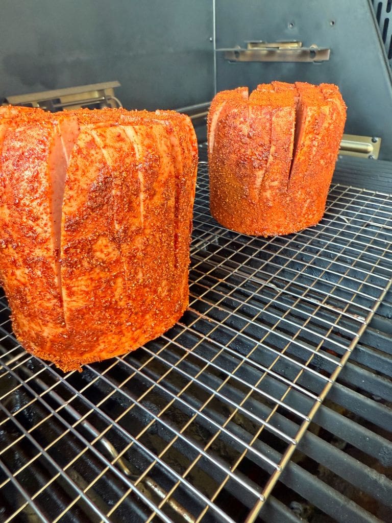 Two large, seasoned cuts of meat are placed on a grill. They are cylindrical, evenly spaced, and covered with a spicy rub. The background shows grill grates, and the scene is part of an outdoor cooking setup.