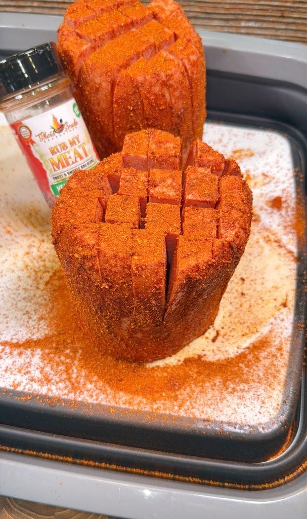 Two seasoned cuts of meat are prepared with a rich, red spice rub, resting on a white tray. A spice jar labeled Rub My Meat is nearby, with spices spread across the surface.
