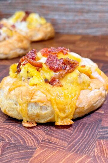 Pastries topped with melted cheese and crispy bacon sit on a wooden surface. The focus is on the pastry in the foreground, showcasing its flaky texture and savory toppings.