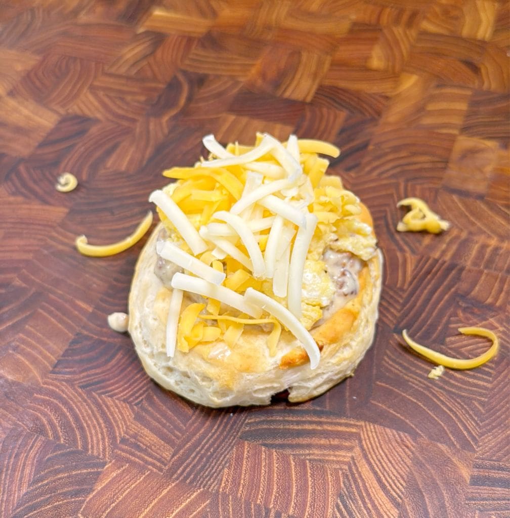 A biscuit topped with scrambled eggs and shredded yellow and white cheese rests on a wooden surface. A few cheese shreds are scattered around it.