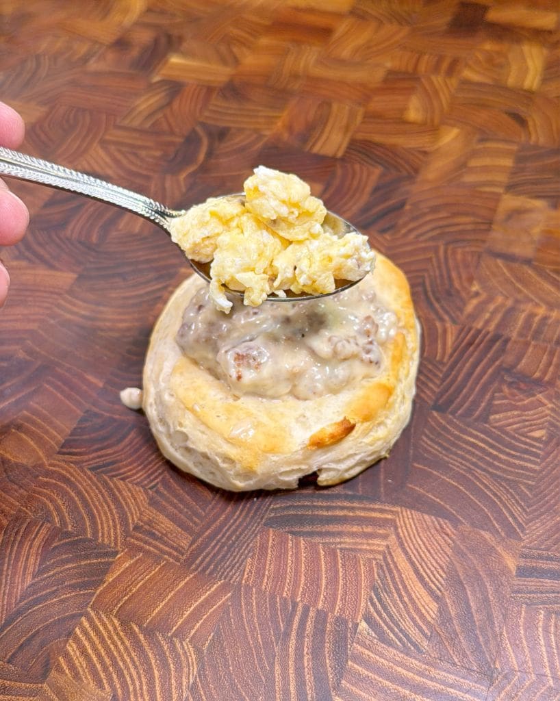 A spoon holds scrambled eggs above a pastry filled with creamy sausage gravy, resting on a textured wooden surface. The pastrys golden, flaky crust contrasts with the smooth gravy and eggs.