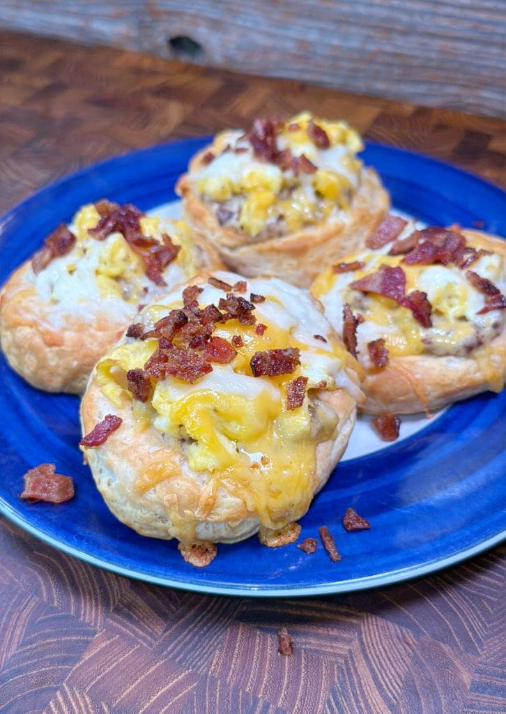 Four savory pastries on a blue plate, topped with scrambled eggs, melted cheese, and bacon bits. The wooden table adds a rustic backdrop.