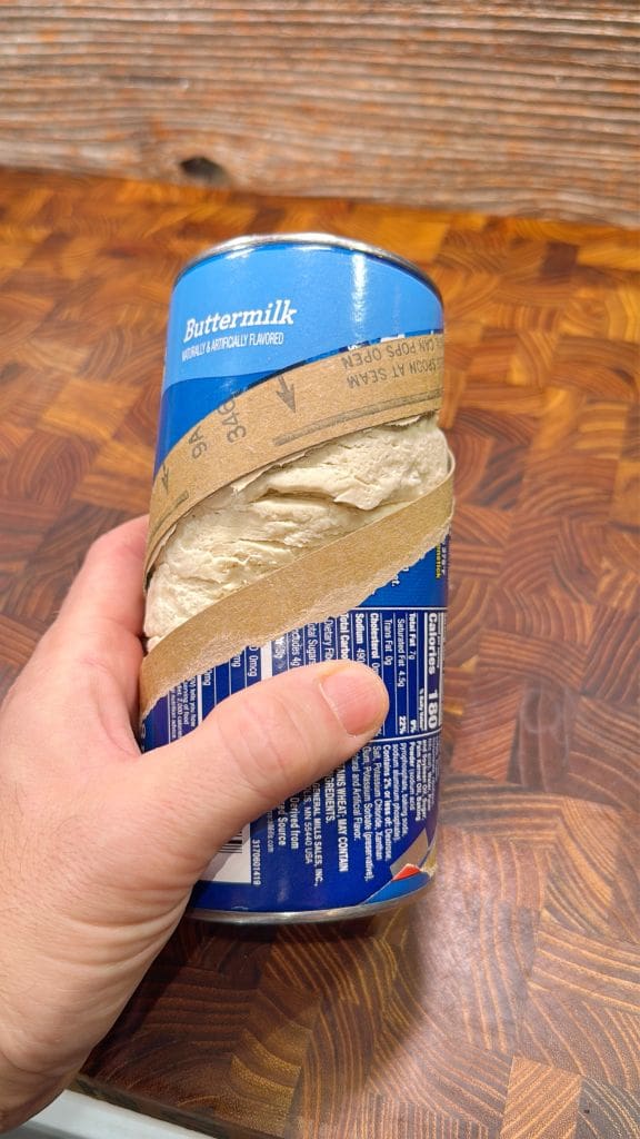 A hand holds a blue canister of buttermilk biscuits, partially opened with the wrapper torn. The canister is on a wooden countertop with a textured, checkered pattern.