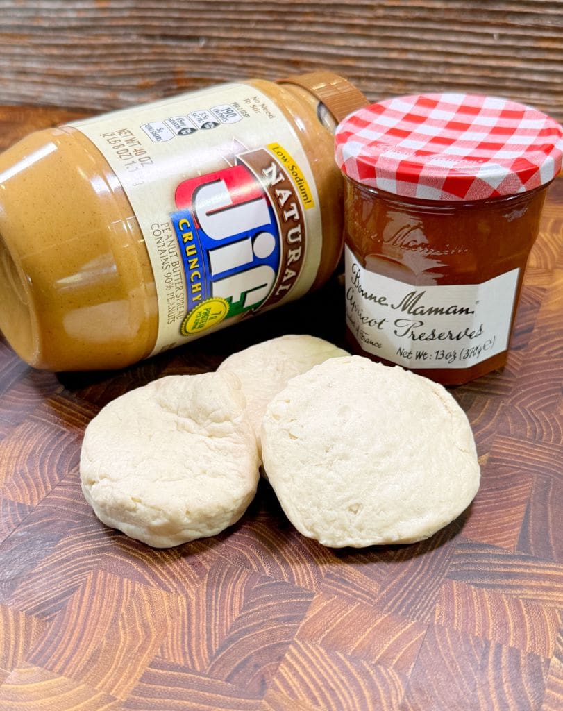 A jar of Jif crunchy peanut butter, a jar of Bonne Maman apricot preserves, and three pieces of biscuit dough are placed on a wooden surface for this homemade uncrustables recipe.