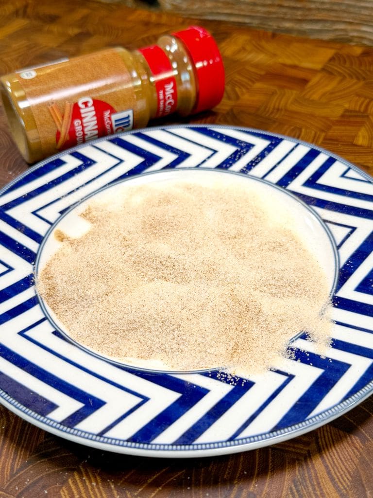 A blue and white patterned plate with a mound of cinnamon sugar powder. In the background, theres a jar of cinnamon sugar with a red lid, tilted on its side on a wooden surface.