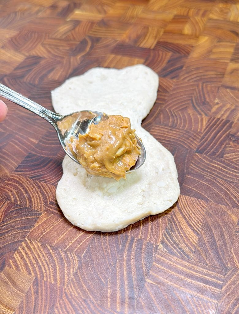 A spoon holds a scoop of peanut butter above a piece of dough on a wooden surface with a herringbone pattern.