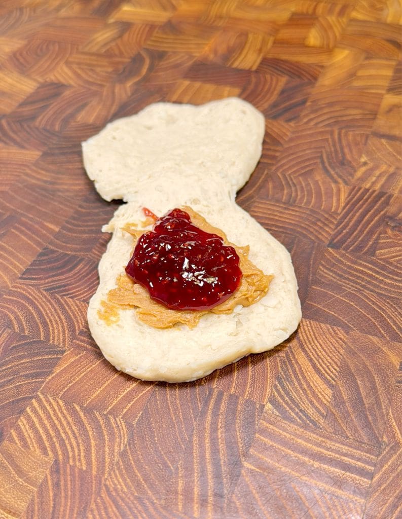 A slice of bread topped with sautéed onions and red jam sits on a wooden cutting board with a swirled pattern. The bread is slightly uneven in shape.