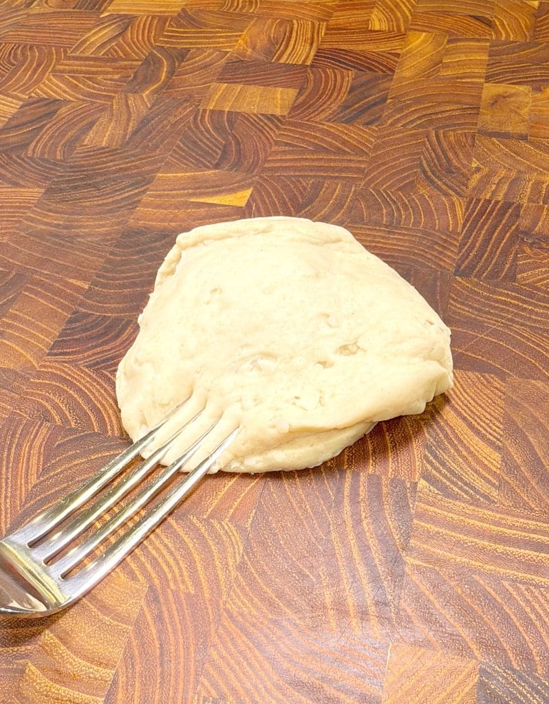 A ball of dough resting on a wooden cutting board with a fork pressing into it slightly. The board features a pattern of various brown shades. The dough appears to be smooth and light in color.