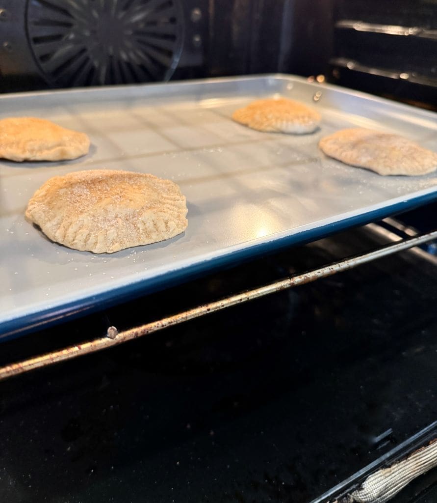 Three unbaked hand pies are on a baking sheet inside an oven. The pies are evenly spaced and the oven door is open, offering a side view of the scene. The ovens interior is dark, highlighting the light-colored pies.