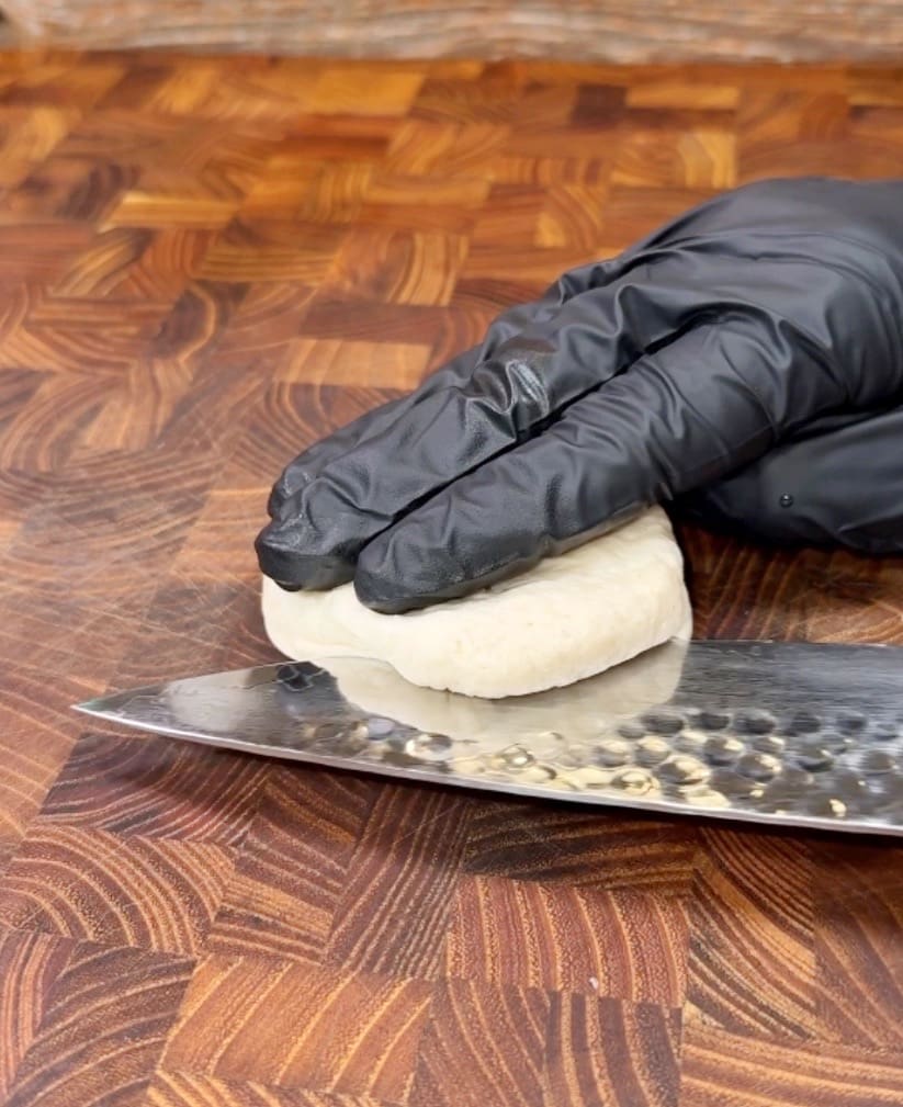 A hand wearing a black glove is pressing down on a small, round dough ball on a wooden cutting board, using a large, textured knife to carefully prepare it for stuffed bombs.