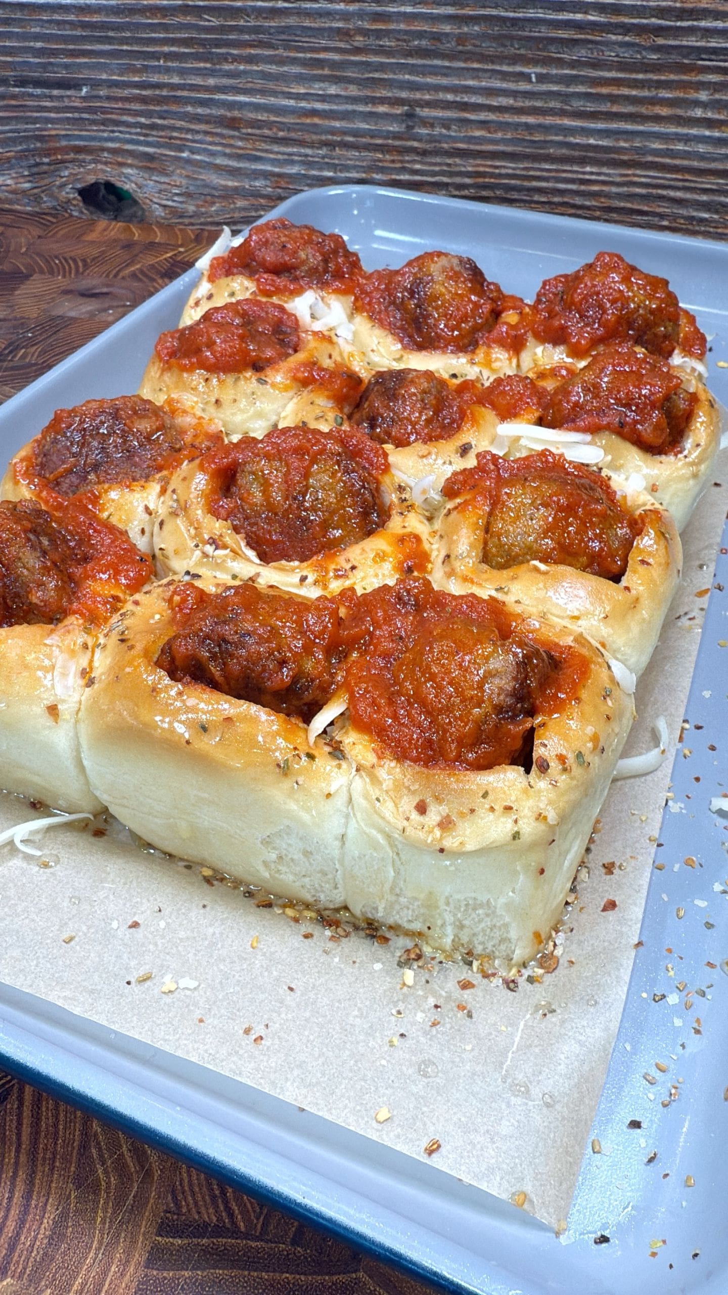 A baking tray with baked bread rolls topped with meatballs and marinara sauce for this meatball sliders recipe. The rolls are nestled closely, with grated cheese and herbs sprinkled on top. The tray is lined with parchment paper and sits on a wooden surface.