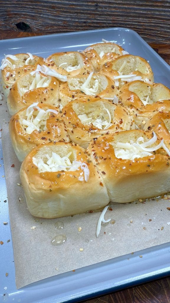 Freshly baked bread rolls topped with melted cheese and sprinkled with herbs and spices on a baking tray lined with parchment paper. The golden-brown crust glistens under the light, and the open woven texture of the rolls is visible.