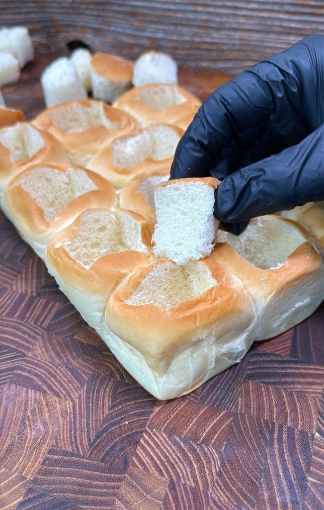 A hand in a black glove lifts a square piece from a batch of dinner rolls. The rolls, set on a wooden surface, are soft and golden-brown, with a piece missing from the center, highlighting the fluffy texture inside.