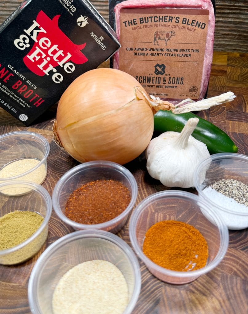 Ingredients on a cutting board: beef broth, a package of beef, a large onion, a garlic bulb, a jalapeño, small containers with various spices including chili powder, paprika, ground pepper, cumin, and onion powder.